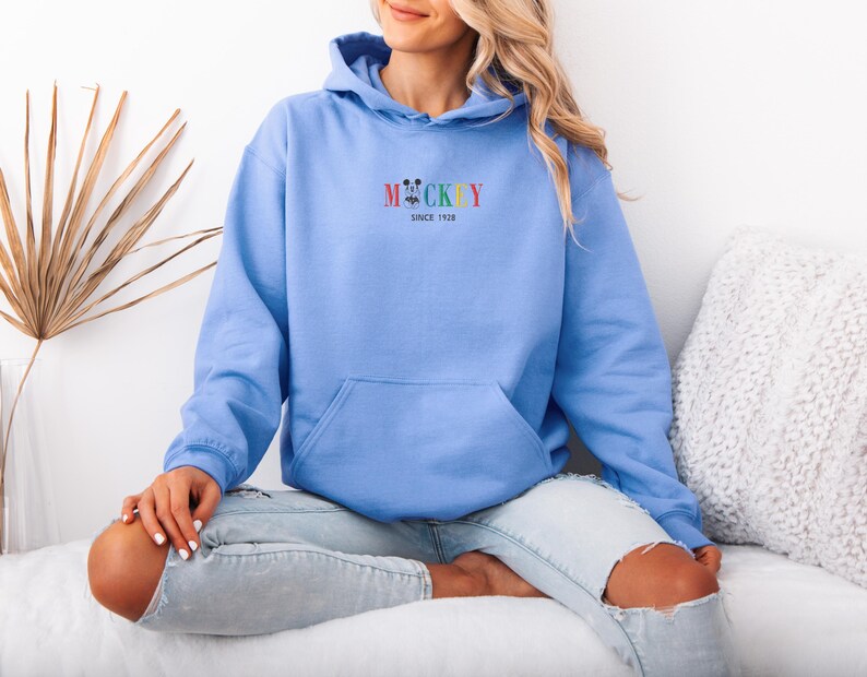 May include: A light blue hoodie with a front pocket and a hood. The word "Mickey" is embroidered in rainbow colors above the text "Since 1928". The hoodie is made of a soft material.