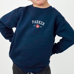Puede incluir: Una sudadera azul marino con el nombre "PARKER" en texto blanco arqueado. Debajo, un peque&ntilde;o c&iacute;rculo rojo con un gr&aacute;fico y el texto "EST. 2001". La sudadera es de cuello redondo y la lleva un ni&ntilde;o.
