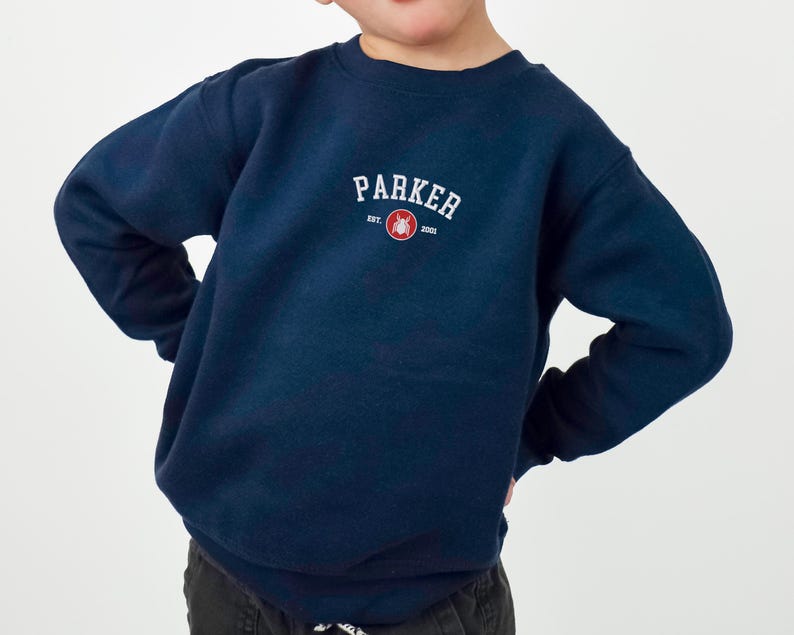 Puede incluir: Una sudadera azul marino con el nombre "PARKER" en texto blanco arqueado. Debajo, un peque&ntilde;o c&iacute;rculo rojo con un gr&aacute;fico y el texto "EST. 2001". La sudadera es de cuello redondo y la lleva un ni&ntilde;o.