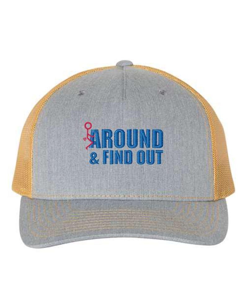 Pu&ograve; includere: Un cappello da camionista grigio e giallo con il testo "AROUND & FIND OUT" ricamato in blu sul davanti. Il testo include un omino stilizzato con il dito medio alzato.