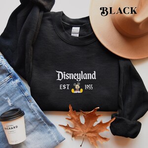 Pu&ograve; includere: Felpa girocollo nera con la scritta "Disneyland" in corsivo bianco, il testo "EST 1955" e una grafica di Topolino. Un cappello color beige &egrave; visibile sullo sfondo. La parola "BLACK" &egrave; stampata nell'angolo in alto a destra.