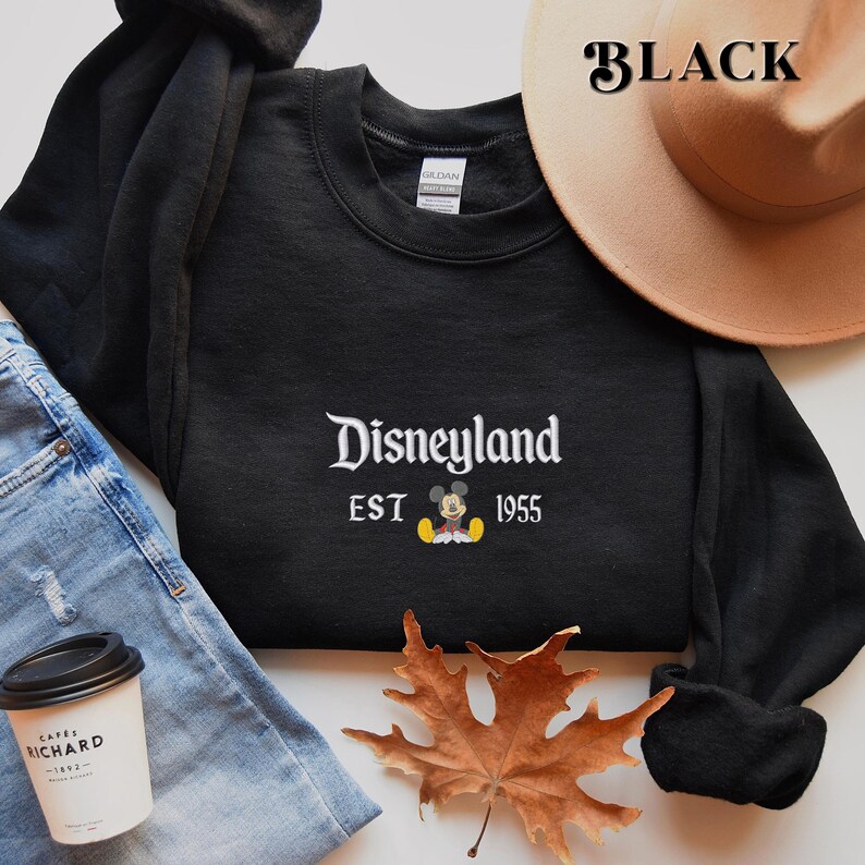 Pu&ograve; includere: Felpa girocollo nera con la scritta "Disneyland" in corsivo bianco, il testo "EST 1955" e una grafica di Topolino. Un cappello color beige &egrave; visibile sullo sfondo. La parola "BLACK" &egrave; stampata nell'angolo in alto a destra.