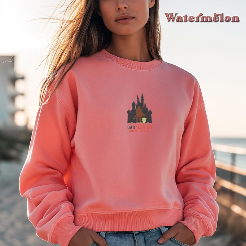 May include: A pink sweatshirt with a graphic of a castle and a figure holding a small green figure. The text "Dadalorian" is below the graphic with the words "This is the way".