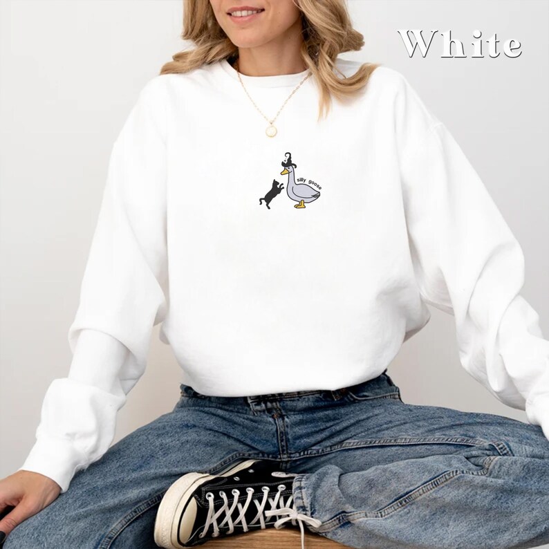 May include: White sweatshirt featuring a black cat and a goose wearing a witch's hat. The goose has the text "Silly Goose" written on it.