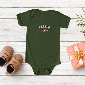 Puede incluir: Un body de beb&eacute; verde oliva con el nombre "PARKER" impreso en texto blanco arqueado. Debajo del nombre hay un gr&aacute;fico rojo y blanco y el texto "EST. 2020". Zapatos de beb&eacute; marrones y un regalo envuelto con un lazo dorado tambi&eacute;n est&aacute;n en la imagen.