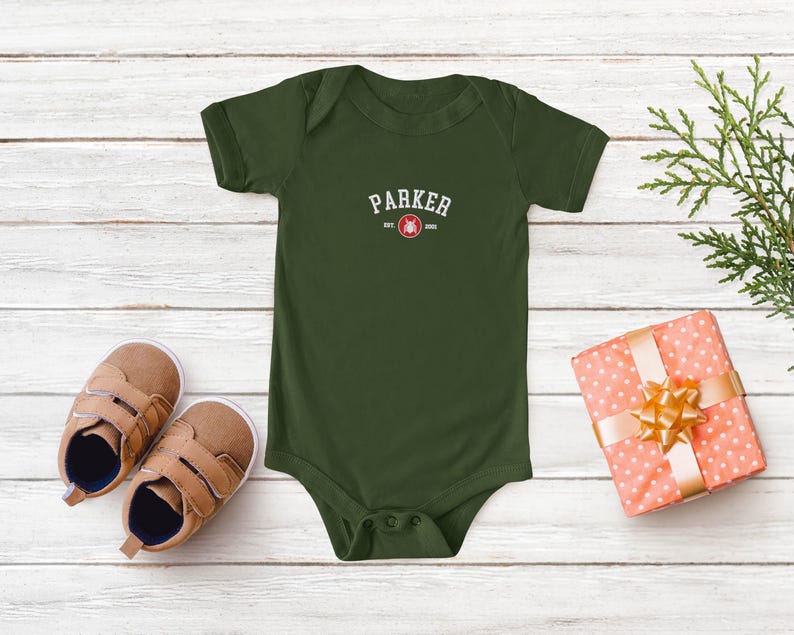 Puede incluir: Un body de beb&eacute; verde oliva con el nombre "PARKER" impreso en texto blanco arqueado. Debajo del nombre hay un gr&aacute;fico rojo y blanco y el texto "EST. 2020". Zapatos de beb&eacute; marrones y un regalo envuelto con un lazo dorado tambi&eacute;n est&aacute;n en la imagen.