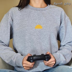 May include: A person wearing a gray sweatshirt with a yellow half-circle design on the front. The person is holding a black video game controller.