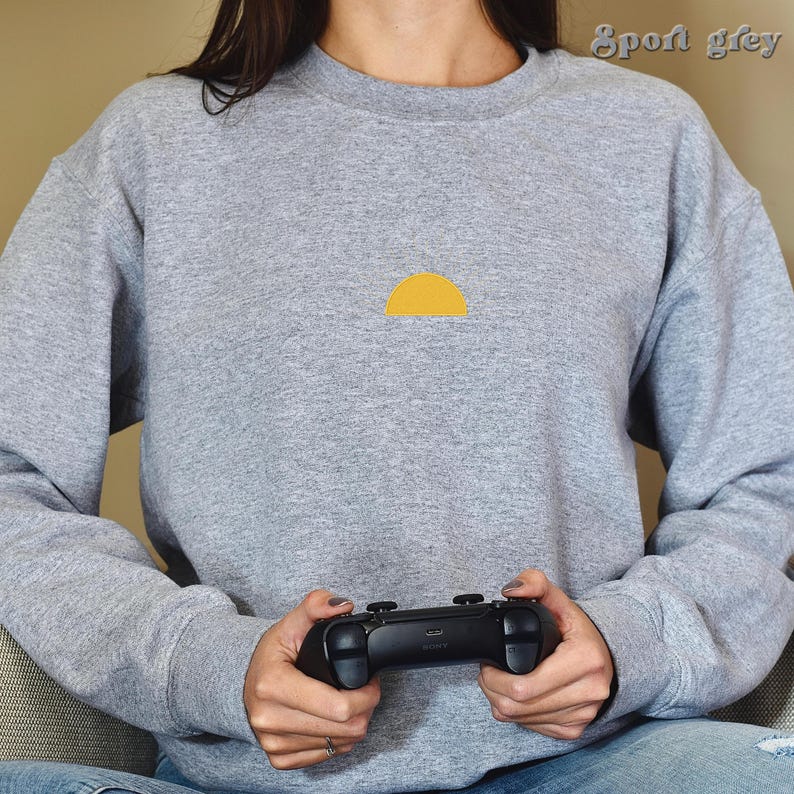 May include: A person wearing a gray sweatshirt with a yellow half-circle design on the front. The person is holding a black video game controller.