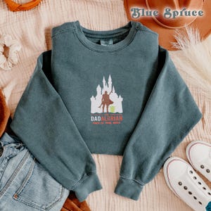 May include: A teal blue sweatshirt with a white silhouette of a castle and a figure holding a green object. The text "The Dadalorian This is the way" is printed below the image.