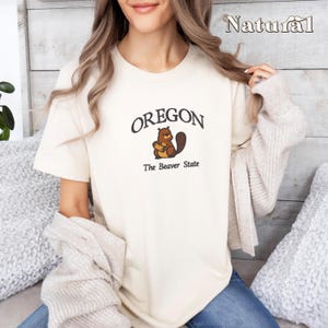 以下が含まれることがあります： 茶色のビーバーのグラフィックと「OREGON The Beaver State」のテキストが書かれた白いTシャツです。