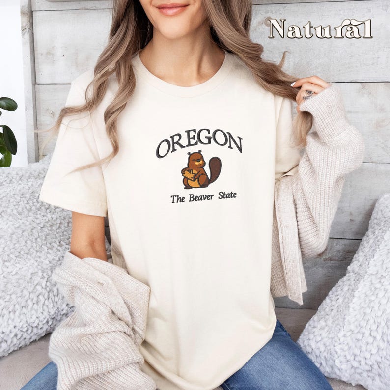 以下が含まれることがあります： 茶色のビーバーのグラフィックと「OREGON The Beaver State」のテキストが書かれた白いTシャツです。