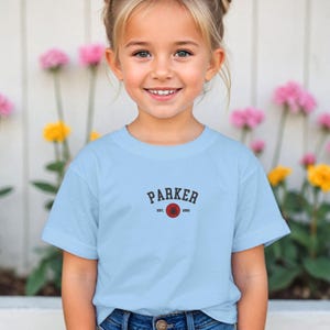 Puede incluir: Una camiseta azul claro con el nombre "PARKER" en letras oscuras sobre un gr&aacute;fico de baloncesto rojo. El texto "EST. 2001" est&aacute; debajo. La camiseta la lleva una ni&ntilde;a.