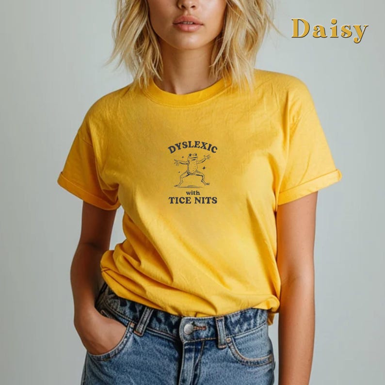 Peut inclure: Un t-shirt jaune avec un graphique noir d'une grenouille et le texte "Dyslexic with Tice Nits".