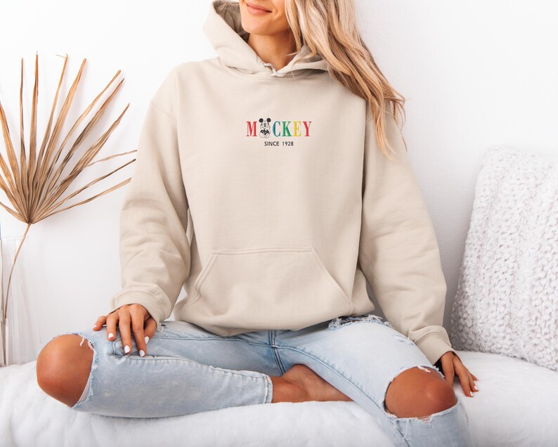 May include: A beige hoodie with a front pocket and a hood. The hoodie features the word "Mickey" in colorful letters with a Mickey Mouse graphic and the text "Since 1928". The person is wearing light blue jeans.