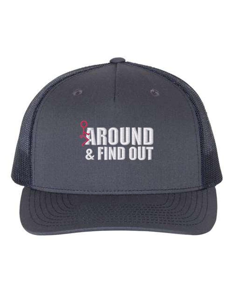 Pu&ograve; includere: Un cappello da camionista blu navy con una rete bianca sul retro. Il cappello ha un design ricamato bianco che dice "AROUND & FIND OUT" con un omino rosso.