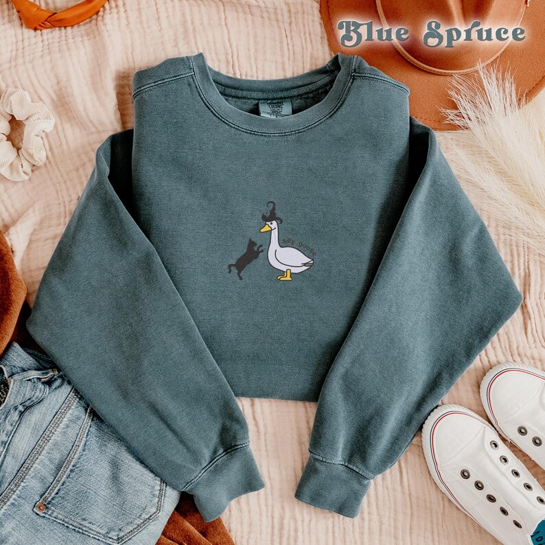 May include: A teal blue sweatshirt featuring a graphic of a goose wearing a witch's hat and a black cat. The text on the graphic reads "Say Woah".