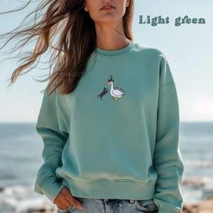 May include: A light green sweatshirt featuring a graphic of a cat chasing a duck wearing a hat. The text "silly goose" is printed on the duck.