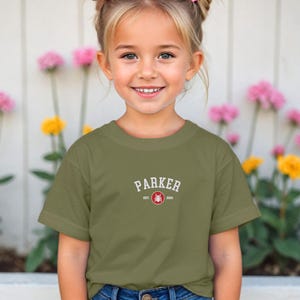 Puede incluir: Una camiseta verde oliva con el nombre "PARKER" en texto blanco arqueado. Debajo, "EST. 2000" y un emblema circular rojo. La camiseta la lleva una ni&ntilde;a frente a una valla blanca y flores.