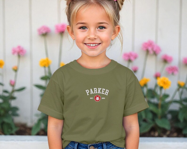 Puede incluir: Una camiseta verde oliva con el nombre "PARKER" en texto blanco arqueado. Debajo, "EST. 2000" y un emblema circular rojo. La camiseta la lleva una ni&ntilde;a frente a una valla blanca y flores.