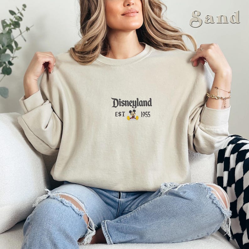 Pu&ograve; includere: Una felpa color sabbia con la scritta "Disneyland" in un font classico, insieme a "EST 1955" e una piccola grafica di Mickey Mouse. La felpa &egrave; a maniche lunghe e ha un girocollo.