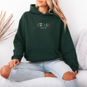May include: Dark green hoodie with a front pocket and a hood. The hoodie features the word "MICKEY" in colorful embroidery, with a small Mickey Mouse head and the text "SINCE 1928" below. The model is wearing light blue jeans.