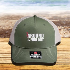 Pu&ograve; includere: Un cappello da camionista verde e bianco con il testo "AROUND & FIND OUT" ricamato sul davanti. Il cappello &egrave; regolabile e ha un logo Richardson sul retro.