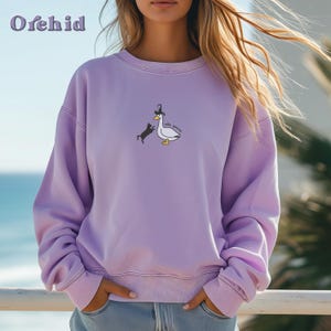 May include: A lavender sweatshirt featuring a graphic of a goose wearing a hat and a black cat chasing it. The text on the graphic reads "silly goose".