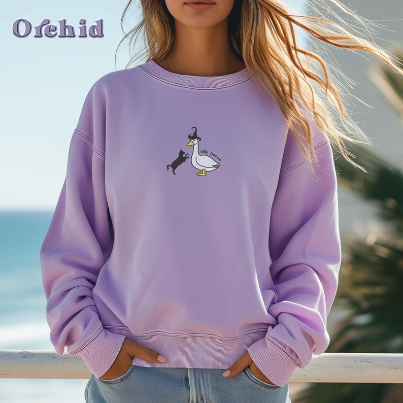 May include: A lavender sweatshirt featuring a graphic of a goose wearing a hat and a black cat chasing it. The text on the graphic reads "silly goose".
