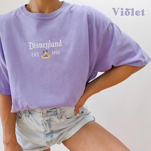 Puede incluir: Una camiseta morada con un gr&aacute;fico blanco de Mickey Mouse y el texto "Disneyland EST 1955".