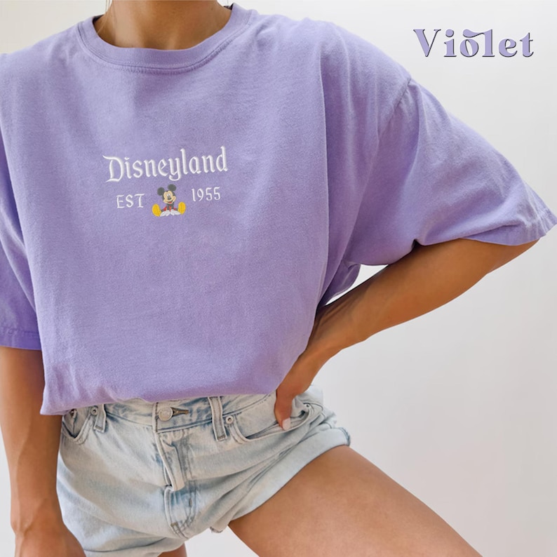 Puede incluir: Una camiseta morada con un gr&aacute;fico blanco de Mickey Mouse y el texto "Disneyland EST 1955".