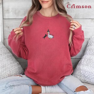 May include: A red sweatshirt featuring a white goose wearing a witch's hat and a black cat with a witch's hat. The text "WITCH PLEASE" is printed on the sweatshirt.