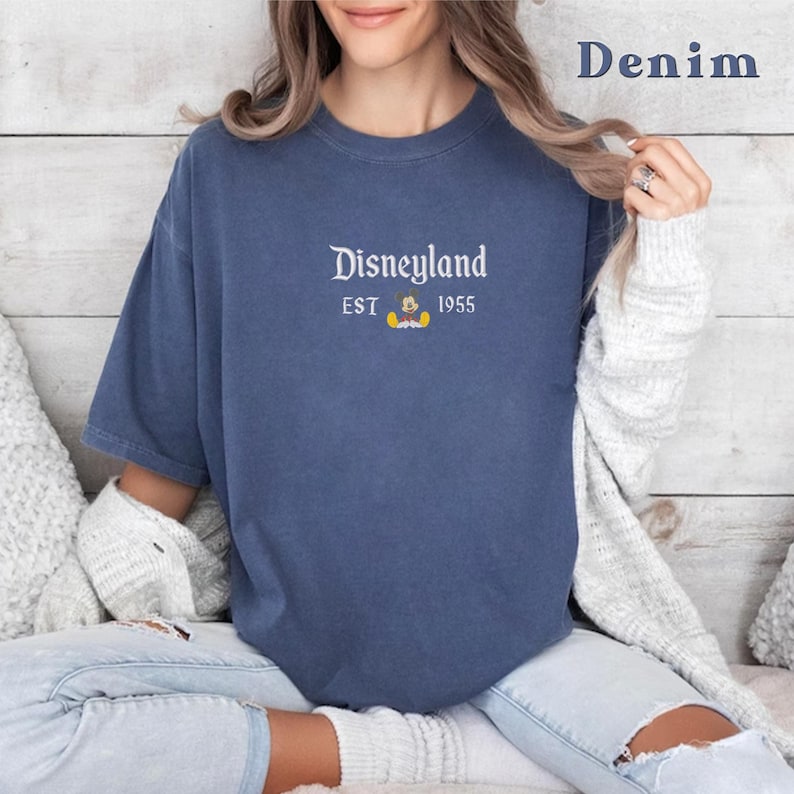 Puede incluir: Una camiseta azul denim con un gr&aacute;fico blanco que dice "Disneyland Est 1955" con un peque&ntilde;o Mickey Mouse.