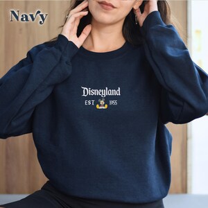 Pu&ograve; includere: Felpa blu navy con la scritta "Disneyland" ricamata in bianco, insieme a "EST 1955" e una grafica di Mickey Mouse. La felpa &egrave; a girocollo.