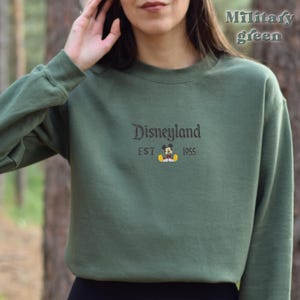 Pu&ograve; includere: Felpa verde militare con la scritta "Disneyland" ricamata in corsivo marrone scuro. Sotto la parola "Disneyland" c'&egrave; la parola "EST" con una grafica di Topolino e il numero "1955".