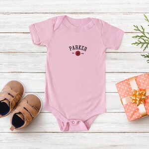 Puede incluir: Un body de beb&eacute; rosa claro con el nombre "PARKER" impreso en rojo oscuro. El body se combina con zapatos de beb&eacute; marrones y un regalo envuelto con un lazo dorado. El fondo es una superficie de madera clara.