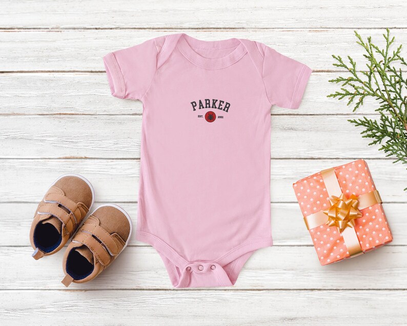Puede incluir: Un body de beb&eacute; rosa claro con el nombre "PARKER" impreso en rojo oscuro. El body se combina con zapatos de beb&eacute; marrones y un regalo envuelto con un lazo dorado. El fondo es una superficie de madera clara.