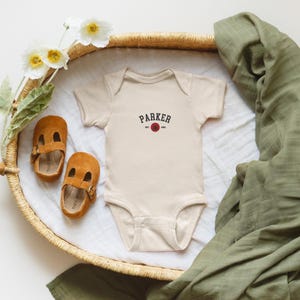Puede incluir: Un body de beb&eacute; beige con el nombre "PARKER" impreso en negro, con un dise&ntilde;o de c&iacute;rculo rojo. Dos zapatos de beb&eacute; de ante marr&oacute;n y una manta de muselina verde tambi&eacute;n est&aacute;n en la imagen. Los art&iacute;culos est&aacute;n colocados en una cesta tejida.