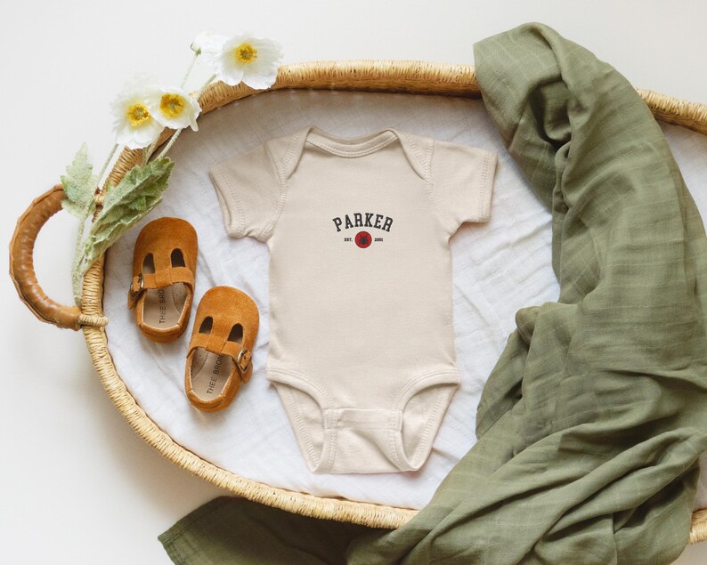 Puede incluir: Un body de beb&eacute; beige con el nombre "PARKER" impreso en negro, con un dise&ntilde;o de c&iacute;rculo rojo. Dos zapatos de beb&eacute; de ante marr&oacute;n y una manta de muselina verde tambi&eacute;n est&aacute;n en la imagen. Los art&iacute;culos est&aacute;n colocados en una cesta tejida.