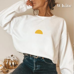 May include: White sweatshirt with a yellow sun graphic. The sun has rays coming out of it. The sweatshirt is being worn by a person.