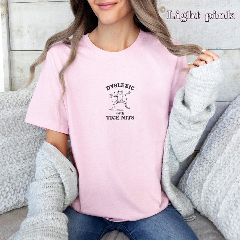 Peut inclure: Un t-shirt rose p&acirc;le avec un graphique d'une grenouille et le texte "Dyslexic with Tice Nits".