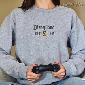 Pu&ograve; includere: Felpa grigio chiaro con la scritta "Disneyland" ricamata in corsivo nero, insieme a "EST 1955" e una piccola grafica di Topolino. La felpa &egrave; uno stile girocollo.