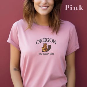 以下が含まれることがあります： ビーバーのグラフィックと「OREGON The Beaver State」のテキストがプリントされたピンクのTシャツです。
