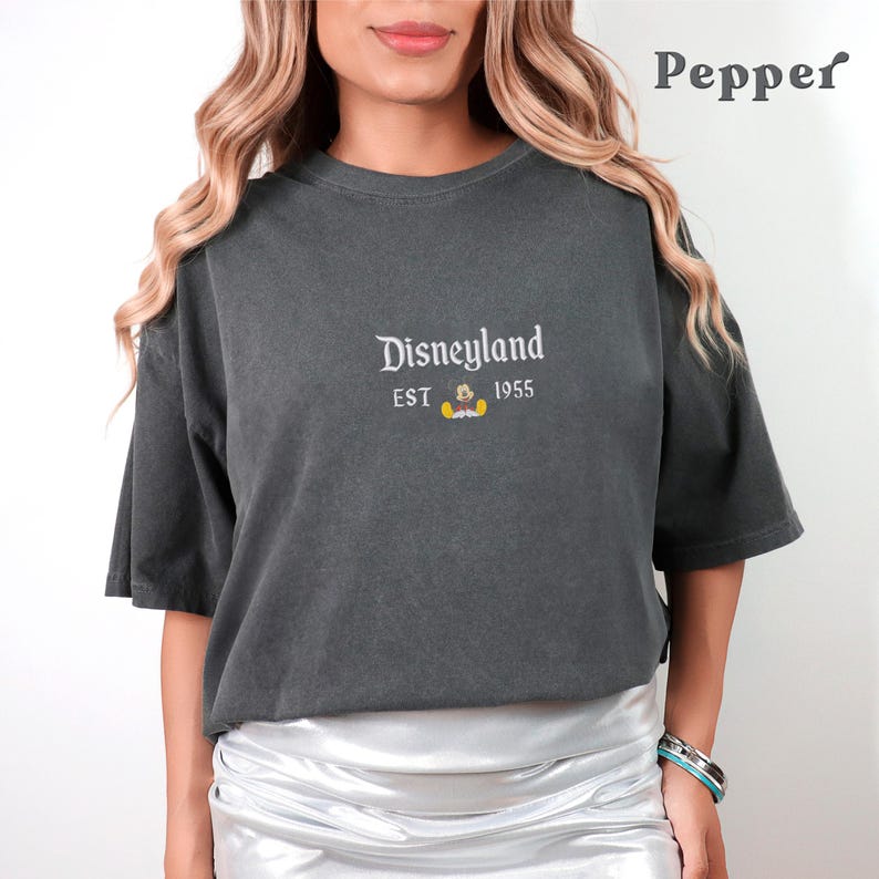 Puede incluir: Una camiseta gris oscuro con un gr&aacute;fico blanco que dice "Disneyland EST 1955" y una peque&ntilde;a imagen de Mickey Mouse. La camiseta la lleva puesta una persona con el pelo largo y casta&ntilde;o.