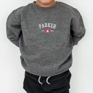Puede incluir: Una sudadera gris oscuro con el nombre "PARKER" en letras blancas arqueadas. Debajo, un dise&ntilde;o rojo y blanco con el a&ntilde;o "2001". La sudadera se combina con pantalones negros.