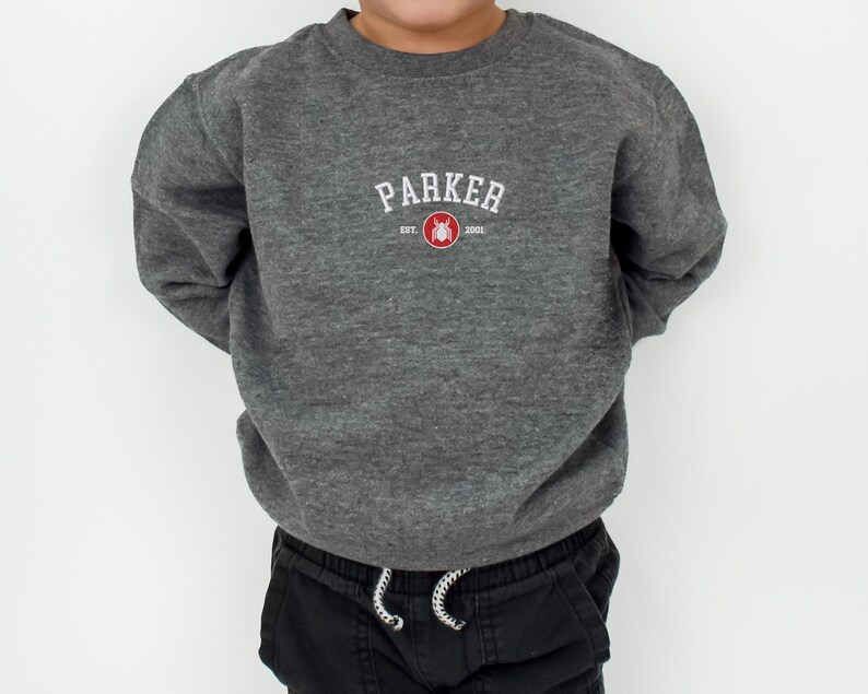 Puede incluir: Una sudadera gris oscuro con el nombre "PARKER" en letras blancas arqueadas. Debajo, un dise&ntilde;o rojo y blanco con el a&ntilde;o "2001". La sudadera se combina con pantalones negros.