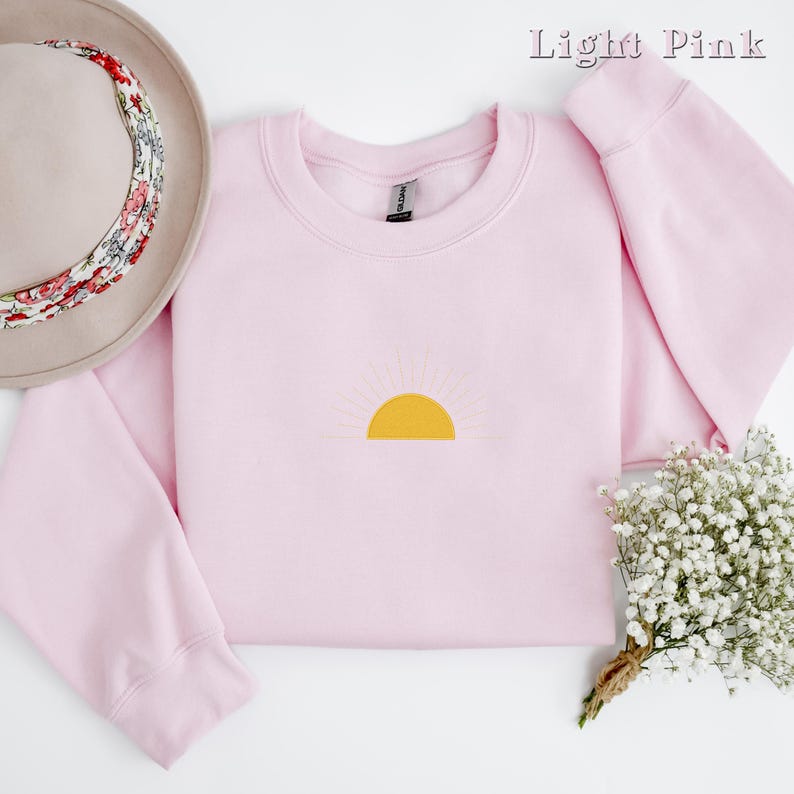 May include: A light pink sweatshirt with a yellow sun graphic. The sweatshirt has a crew neck and long sleeves.