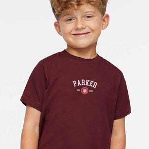 Puede incluir: Una camiseta burdeos de manga corta con la palabra "PARKER" en texto blanco arqueado. Debajo del texto hay un peque&ntilde;o gr&aacute;fico y el texto "EST. 2001". La camiseta est&aacute; hecha de un material suave.