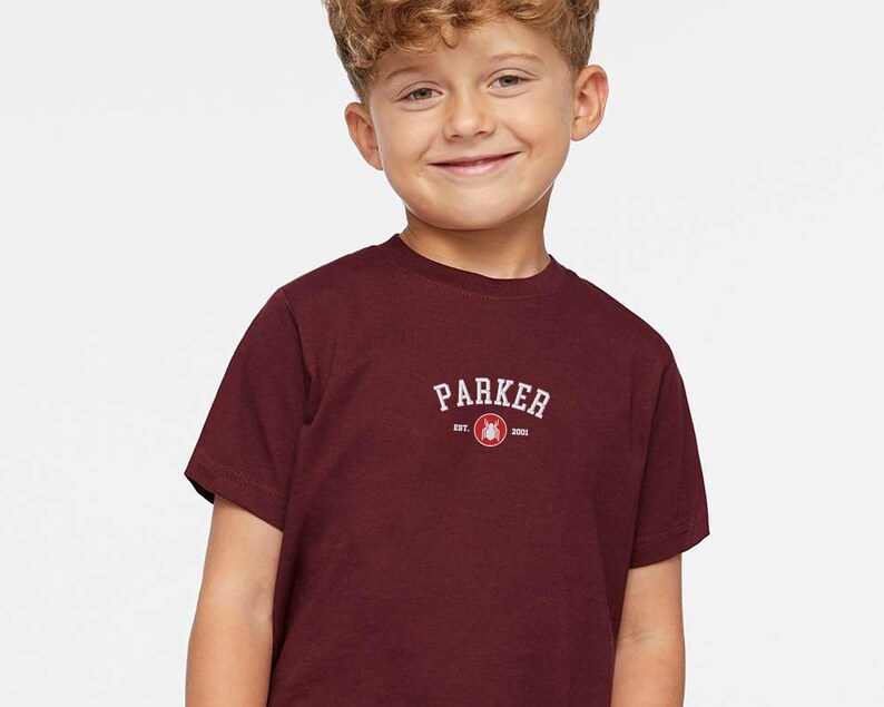 Puede incluir: Una camiseta burdeos de manga corta con la palabra "PARKER" en texto blanco arqueado. Debajo del texto hay un peque&ntilde;o gr&aacute;fico y el texto "EST. 2001". La camiseta est&aacute; hecha de un material suave.