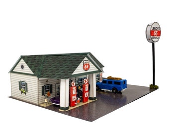 May include: A model of a vintage gas station with a white building, green roof, and red Route 66 sign. The gas station has two red gas pumps and a blue car parked in front. A sign with the text "Leroy's 66 Service" is on a pole in the background.