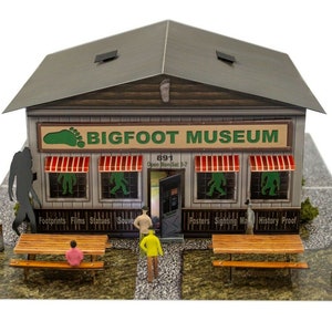 May include: A miniature model of a Bigfoot Museum building with a brown roof, gray walls, and red-striped awnings over windows. The sign reads Bigfoot Museum 891 Open Mon-Sat 9-7. Small figures and picnic tables are placed in front of the structure.